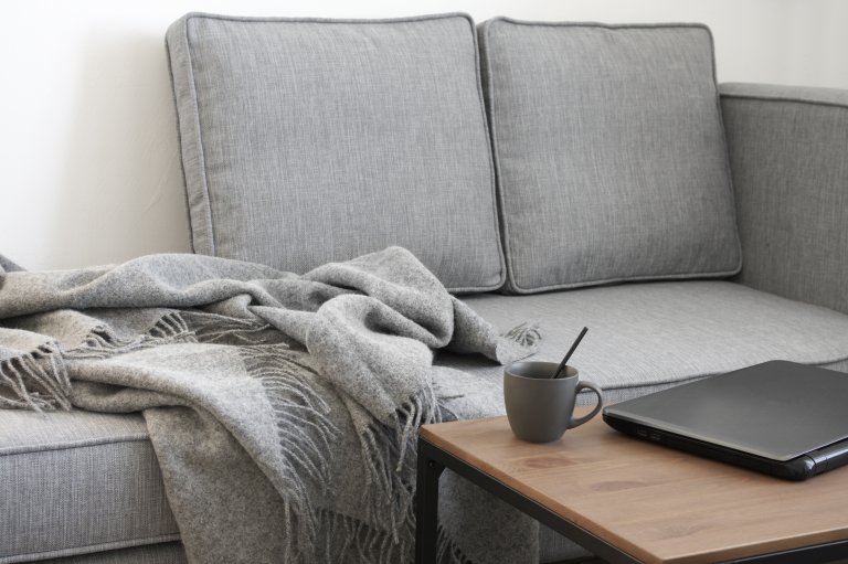 A cozy gray sofa with a matching throw blanket draped over one arm is positioned against a white wall, creating a comfortable workspace. A wooden coffee table in front of the sofa holds a mug with a spoon and a closed black laptop.