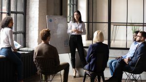 A woman stands presenting to a group of five people in a modern office setting. She uses a whiteboard with diagrams, exuding the confidence of a boss. The group is seated in a semi-circle around her, attentively listening. Large windows and an industrial-style interior provide natural light.