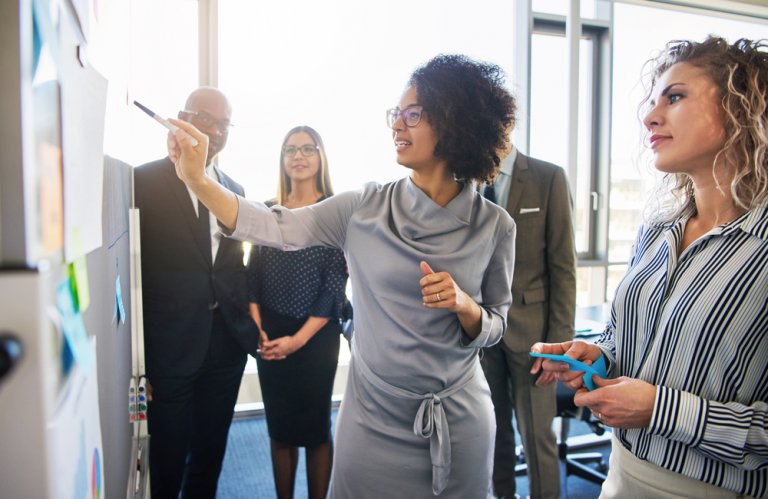 A woman presenting at a whiteboard in a modern office setting, pointing with a marker while two women and a man look on attentively. They all appear engaged and focused on the presentation, showcasing the collaborative spirit of the women workforce in this bright, contemporary environment.
