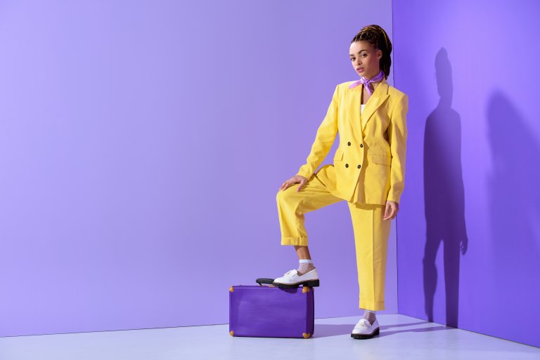 A woman in a bright yellow suit and white shoes poses confidently with one foot on a purple suitcase against a matching purple background. Her braided hair and serious expression capture the essence of current fashion trends, creating a vibrant and striking contrast within the minimalist setting.