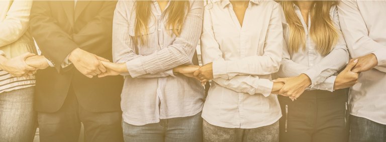 A line of people standing side by side, holding each other's arms in solidarity and a sense of belonging. The group consists of both men and women wearing casual and business attire. The background has a warm, sunlit glow.