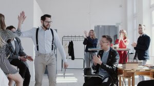 A smiling man with glasses and suspenders gives a high five to a woman in a bright company office. Around them, colleagues stand and sit, clapping and cheering. The office has large windows, a clothes rack, and tables with various items. Everyone appears joyful.