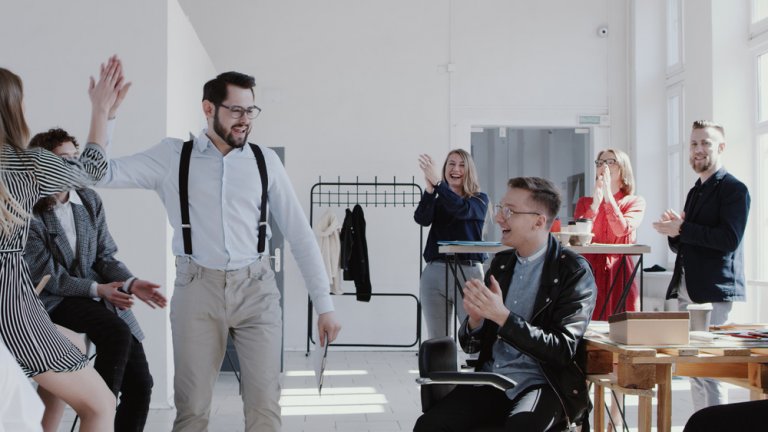 A smiling man with glasses and suspenders gives a high five to a woman in a bright company office. Around them, colleagues stand and sit, clapping and cheering. The office has large windows, a clothes rack, and tables with various items. Everyone appears joyful.