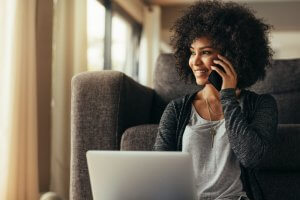A person with curly hair is smiling while talking on a smartphone. They are sitting on the floor next to a couch, with a laptop on their lap, creating an impromptu work space. The setting appears to be a well-lit living room with large windows in the background.