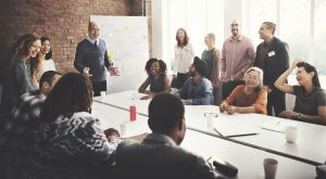 A diverse group of people in a meeting room are gathered around a long table, with the boss at the head. Some are standing, while others are seated. They appear to be engaged and smiling. A whiteboard with notes and diagrams is visible in the background, all well-lit by natural light.