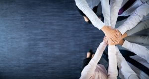 A group of people, seen from above, place their hands together in a display of teamwork and unity, fostering a sense of belonging. They are standing on a dark blue carpeted floor, wearing business attire, with only the upper halves of their bodies visible in the frame.