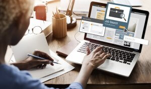 A person sits at a desk, typing on a laptop with a "Job Search" website open. Various office supplies, including a notebook, pen, and smartphone, are scattered on the desk. A desk light and a cup are visible in the background as they focus intently on their job hunt.