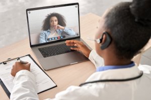A doctor wearing a white coat and headset sits at a desk, conducting a video call on a laptop with a young woman. The doctor holds a clipboard with a form as they discuss mental health. The woman appears to be seated at home, engaged in the virtual consultation.
