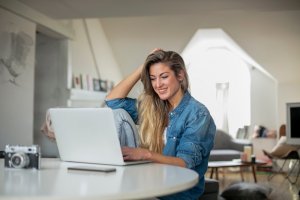 A woman with long blonde hair sits at a white table in her work space, using a laptop. She is wearing a blue denim shirt and smiling while looking at the screen. The background shows a bright room with modern décor, including a bookshelf, camera, and furniture.