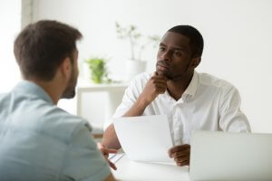 Two men are seated across from each other at a table, engaged in conversation. The boss, holding a document, is attentively listening with a contemplative expression while the other man speaks. A laptop and plants are visible in the background.