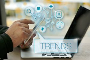 A person holds a smartphone in their left hand while using a laptop on a desk. Various technology icons, including a magnifying glass, calendar, and email, float above the devices. The word "TRENDS" prominently highlights the latest tech trends in the foreground.