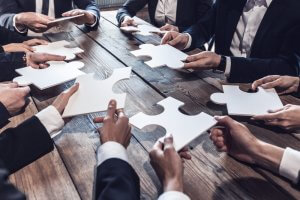 A group of people in business attire, sitting around a wooden table, each holding a large white puzzle piece. The puzzle pieces are held close together, suggesting a collaborative effort to fit them together and a strong sense of belonging.