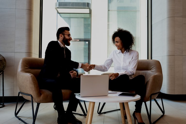 A man and a woman, both in business attire, sit across from each other on beige chairs in an office setting, shaking hands over a table with a laptop on it. They appear to be concluding a successful meeting about careers in government. Large windows in the background let in ample light.