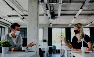 Two people wearing face masks sit at separate desks in a modern office space with exposed pipes and concrete walls. They are engaged in a conversation about careers in government, gesturing with their hands, as natural light streams in through large windows in the background.