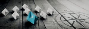 A group of paper boats, with an outlier blue boat leading white boats on weathered wooden planks, mirrors the journey toward government jobs. A large compass design is overlaid on the right side of the image, symbolizing direction and leadership. The scene is in partial grayscale.
