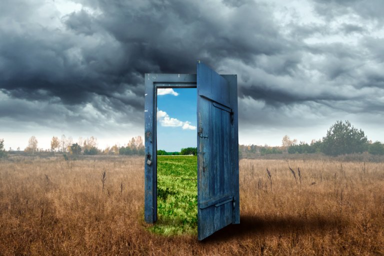 A weathered blue door stands open in the middle of a dry, brown field under a dark, stormy sky. Through the door is a bright, sunny landscape with green grass and a clear blue sky, much like careers in government offering hopeful opportunities amid challenging times.
