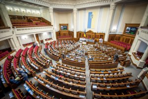 A large, semi-circular parliamentary chamber with rows of wooden desks and red chairs arranged in concentric arcs. The room has high ceilings, tall columns, and a national flag displayed prominently at the front. Several people are seated and engaged in discussions about careers in government.