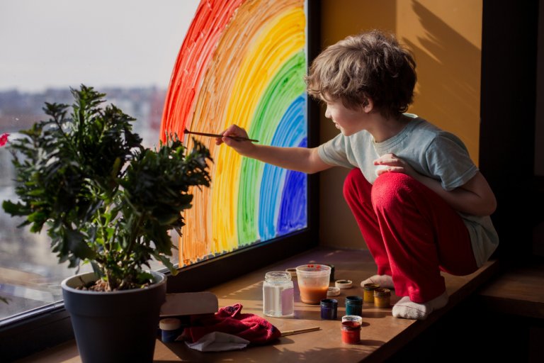 A young child with curly hair, wearing a light blue shirt and red pants, is kneeling on a window seat painting a colorful rainbow on the window. Beside them sits a potted plant along with paints and brushes. The outside view is blurred, reflecting the windows of opportunity in careers in government.