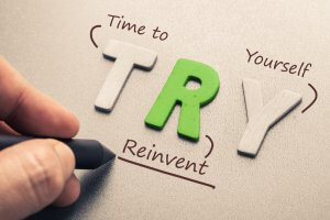 A hand is holding a pen, circling the letters T, R, and Y, spelled out in block letters on a textured surface. The text surrounding the letters reads: "Time to Reinvent Yourself." The T and Y are in white, and the R is in green. Perhaps it's time to explore careers in government for your next step.