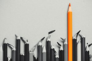 A row of mostly broken or damaged gray pencils lies on a light gray background, with one intact and sharpened orange pencil standing upright and taller than the rest, drawing attention—much like those who excel in Government Jobs.