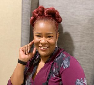 A smiling woman with reddish-brown hair styled in intricate buns, wearing a purple floral-patterned top. She is sitting against a gray upholstered backdrop and resting her hand near her face, perhaps contemplating a future in government jobs.