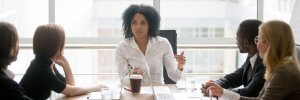 A woman with curly hair, dressed in a white shirt, leads a meeting at a conference table. Four colleagues, three men and one woman, listen attentively as they discuss careers in government. The table has documents, a laptop, and a coffee cup. Large windows are in the background.