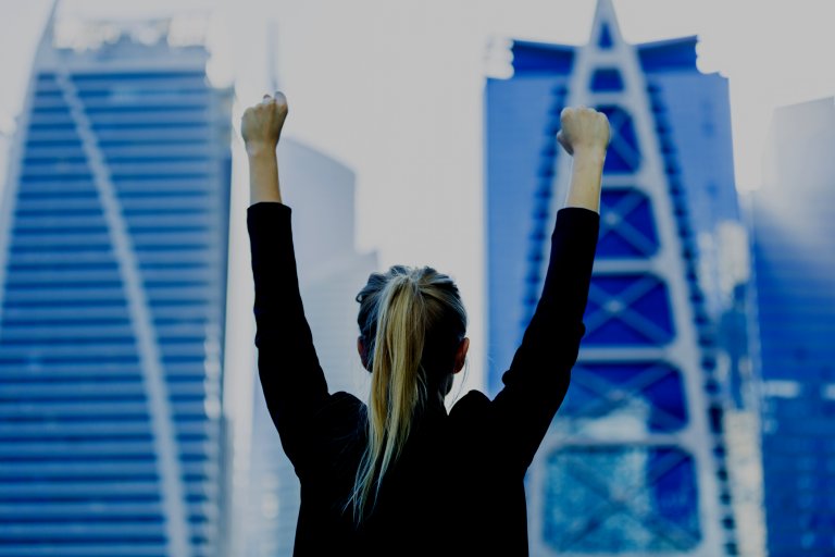 A person with long blonde hair in a ponytail stands with both arms raised triumphantly in front of a cityscape with modern skyscrapers, celebrating their new career in government. The person wears a black jacket, facing away from the camera, and the blue sky contrasts with the buildings in the background.