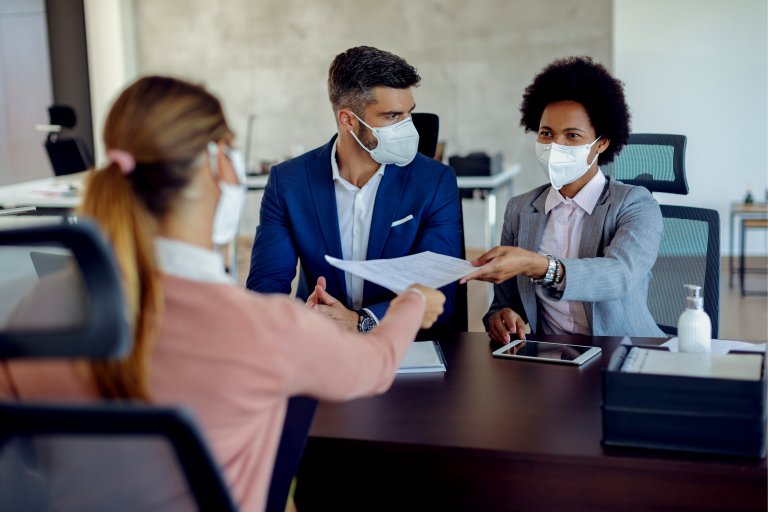 Three colleagues, wearing face masks, sit at a table in a modern office. One person is handing a document to another while the third person looks on. A tablet and hand sanitizer are on the table, highlighting the careful measures even in discussions about careers in government.