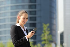 A woman in a black blazer and white shirt is outdoors, smiling while looking at her phone. She stands against the background of a modern glass building and greenery, suggesting an urban setting. Could she be exploring careers in government?