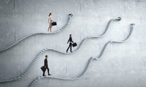 Three business professionals, two women and one man, carrying briefcases, ascend three separate, winding concrete staircases against a gray wall. Each staircase goes in different directions, symbolizing various Government Jobs or challenges within careers in government.