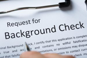 A clipboard holds a form titled "Request for Background Check." A hand is partially visible, holding a pen as if filling out the form. The form includes a certification statement related to the accuracy and honesty of the provided information, essential for careers in government.