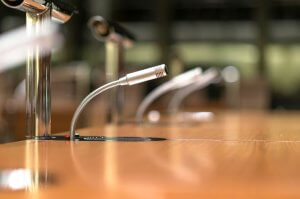 A close-up of several microphones on flexible stands installed on a polished wooden surface, likely in a conference room or meeting hall where discussions about government jobs are held. The microphones are positioned in a line, extending towards the center of the room. The background is blurred.