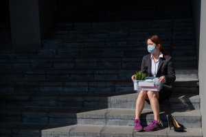 A woman in a business suit and face mask sits on outdoor steps, holding a box with office supplies, paperwork, and a potted plant. Her high heels are off to the side, and she is wearing purple sneakers. The scene suggests job loss or resignation from her Government Job.
