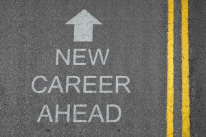 A road marked with double yellow lines on the right side. Painted on the asphalt are the words "NEW CAREER AHEAD" accompanied by a large white arrow pointing straight ahead, highlighting opportunities in careers in government.