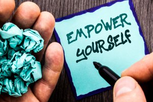 A hand holds a crumpled piece of turquoise paper, while another hand writes "EMPOWER YOURSELF" in bold letters on a turquoise sticky note with a black marker. The background is a dark wood surface, subtly hinting at the stability and promise found in Government Jobs.