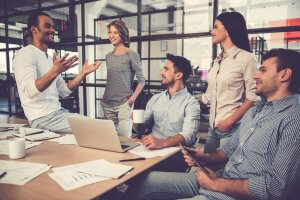 A group of five people, three men and two women, are having a casual meeting in a modern office. One man is standing and speaking animatedly about careers in government, while the others sit around a table with a laptop, documents, and coffee cups, listening and smiling.