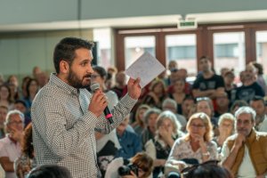 A man with a beard and short hair is speaking into a microphone while holding a piece of paper. He is standing in front of a large audience seated indoors, discussing careers in government, with a mix of ages and expressions among them. The background shows windows and a door.