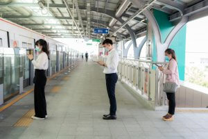 Three people wearing masks stand in line on a train platform, maintaining social distance. They are focused on their phones while waiting for the train to arrive, possibly browsing listings for government jobs. The platform is covered, and the train is partially visible on the left.