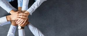 A group of seven people join hands in the center of the image, symbolizing teamwork and unity. They are wearing business attire, representing their dedication to careers in government, with a dark grey textured surface in the background.