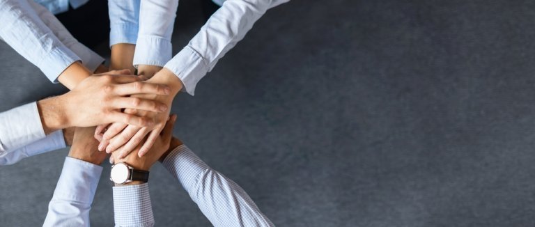 A group of seven people join hands in the center of the image, symbolizing teamwork and unity. They are wearing business attire, representing their dedication to careers in government, with a dark grey textured surface in the background.