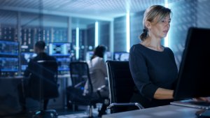 A woman in a dark dress works intently on a desktop computer in a dimly lit office filled with monitors and servers. Two other people, partially obscured, are seated at desks in the background, also working on computers. The atmosphere is high-tech and focused, typical of careers in government.