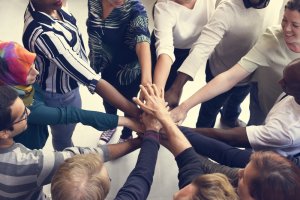 A diverse group of individuals stands in a circle, each person reaching in to stack their hands together at the center, symbolizing unity and teamwork. They display various expressions of positivity and cooperation, united by their commitment to meaningful careers in government.