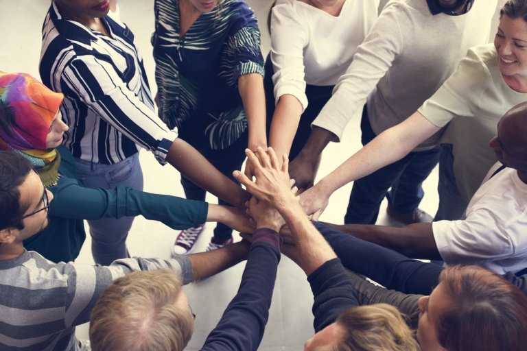 A diverse group of individuals stands in a circle, each person reaching in to stack their hands together at the center, symbolizing unity and teamwork. They display various expressions of positivity and cooperation, united by their commitment to meaningful careers in government.