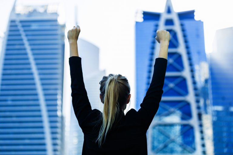 A person with long blonde hair in a ponytail, wearing a black jacket, stands with their back to the camera and their arms raised in triumph. This confident leader faces several modern high-rise buildings in the background.