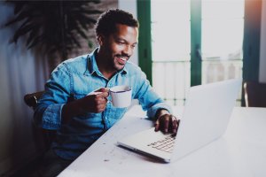 A man sits at a table, smiling and holding a white mug in his right hand while typing on a laptop with his left. He is wearing a light denim shirt and appears to be researching careers in government. The bright room with large windows in the background adds to the serene atmosphere.