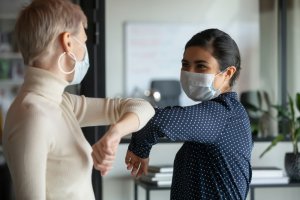 Two women wearing face masks greet each other with an elbow bump in an office setting. One woman, with short blonde hair and a white shirt, is engaged in a conversation about careers in government with the other woman, who has dark hair tied back and is dressed in a navy blue polka-dot shirt.