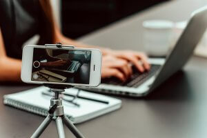 A smartphone is set up on a tripod, recording the hands of a person typing on a laptop. A notebook and pen are placed nearby on the desk, suggesting they're researching careers in government. In the background, a blurred coffee cup is visible, emphasizing a focused work or study environment.