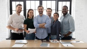 A group of six smiling people, led by a confident leader, stands together in a well-lit office with large windows. They are dressed casually, with three holding tablets or papers. A wooden table with laptops, documents, and glasses of water is in front of them.