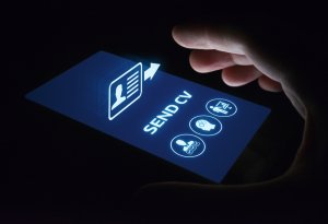 A glowing, backlit smartphone screen displays a "SEND CV" button with an icon of a profile and arrow. Beneath it are icons for a briefcase, chat bubble, and people. A hand hovers over the screen, preparing to tap it against a dark background—ready to explore careers in government.