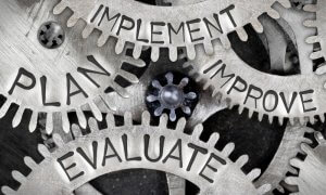 Close-up of interlocking metal gears with words on them: "PLAN," "IMPLEMENT," "IMPROVE," and "EVALUATE." The gears convey the interconnectedness and cyclical nature of these processes, much like careers in government where each role is integral to making a difference.